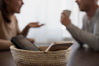 Two phones in a basket on a table in front of a man and woman talking.