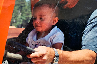 An image of me at 3, on my dad’s lap, driving an orange forklift with trees in the background.