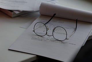 pair of glasses laying on a pad of paper with writing on it — you should proofread your Medium stories.