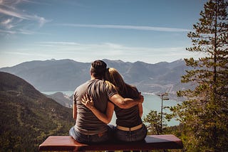 Man and woman sit on bench overlooking beautiful mountain range and lake.