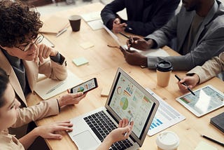 Grupo de pessoas em uma reunião ao redor de uma mesa de madeira clara. Na mesa, há diversos dispositivos tecnológicos, como laptops, tablets e smartphones, exibindo gráficos e dados. Há também papéis, canecas de café, e canetas espalhadas pela mesa.