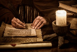 Close up of a man’s hands writing on parchment in a dark room with candle and old books piled up nearby.
