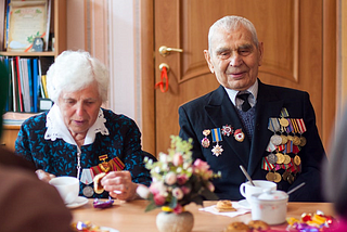 Photo of an eldery lady and eldery man sitting at a table with coffee. The old man is a war veteran wearing a suit ocvered in medals