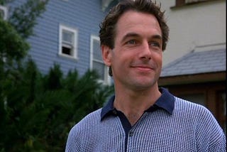 A color image from The Deliberate Stranger, showing Mark Harmon with a smile as he portrays the charming side of Ted Bundy. He has tussled dark hair and blue eyes, and he is wearing a shirt with a dark blue collar and a blue and white pattern. Houses are visible behind him.