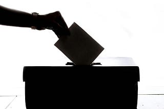 A black on white image of a hand placing a vote into a ballot box.