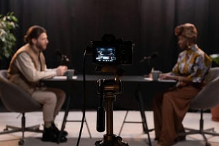 A camera is aimed at two people sitting at a table. Microphones are placed on the table in front of them.