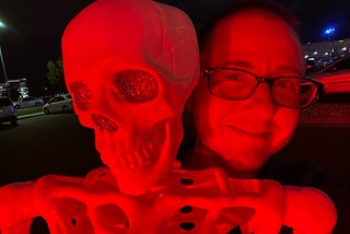 The author is standing behind a skeleton under a red light. He has very short hair and glasses. He’s smiling. The skeleton’s eyes are decorated with glitter.