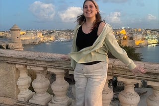 A girl wearing a white pants, a black shirt tank top and a light green overshirt stands on a bridge overlooking a city landscape.