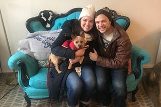Megan and Tim pose happily with their new dog, Arthur, a Cairn terrier-chihuahua mix, on a blue couch.
