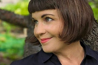 A lady with brown hair and red lipstick in front of a tree smiling