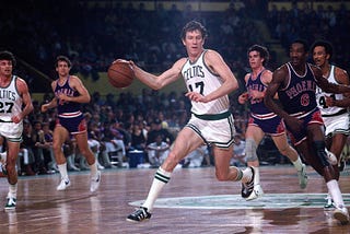 A scene from a historic NBA game featuring intense basketball action between the Boston Celtics and Pheonix Suns in the 1976 NBA Finals. Players in basketball jerseys are seen running, dribbling, and standing still. The image captures the excitement of the game with players engaged in intense competition.