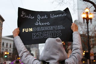 Figure in hoodie holding up a “Girls just wanna have fundamental rights” sign