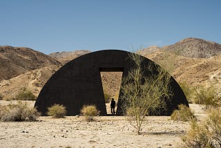 A giant, half-moon, black sculpture sits in the desert landscape. Behind it are sandy hillsides. At its center is an angular cutout, a doorway with the silhouette of a person standing inside.