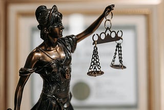 a close-up photo of a bronze figurine of Lady Justice from the waist up, with a blurred-out academic degree in a picture frame in the background
