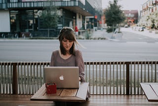 A women on her mac laptop facing the camera, sitting in a open patio cafe looking on to the intersection
