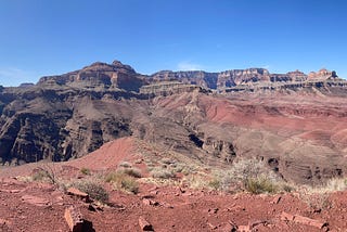 Panorama photo of the Grand Canyon. Blue sky above red rocky soil with a river at the bottom of a deep canyon. There is a shadow of a hiker on the right side.