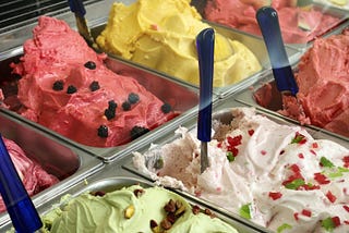 A traditional gelato display with several different flavours in metal trays with spatulas stuck into the,