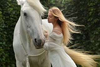 A woman with blonde hair standing next to a big white horse