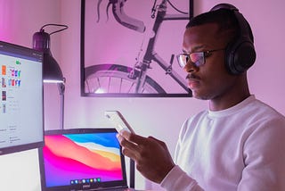 Man with eyeglasses and headset wearing white shirt looks at his phone and sits in front of a laptop and monitor