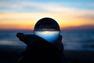 Shadow hand holds a crystal ball up to the ocean sunset so it shows up inverts in the ball.