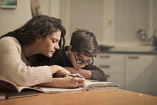 Mom leaning over and helping son who is writing in a book at their kitchen table.