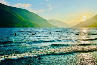 A shoreline with waves rolling in. Children play in the water further out. The sun sets behind hills in the distance