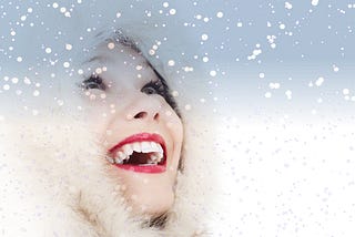 A young woman wearing a fur-trimmed hood looks up into falling snow, laughing.