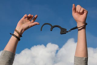 A photograph shows a person’s forearms raised up. Gray shirtsleeves are barely visible at the bottom of the image. The person has a woven or beaded multicolor bracelet and a dark colored leather bracelet on their right wrist. They have one locked handcuff on their left wrist. Attached is an open handcuff that swings in the air between their hands. A blue sky with puffy white clouds is shown in the background.