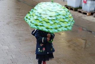 A toddler walking on a wet pavement, her face obscured by a large green umbrella