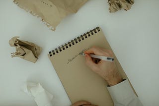 A hand is shown, holding a pen above a notebook with an entire sentence scratched thorough. There are 2 crumpled pieces of paper, indicating the author in the picture was not happy with previous writing attempts.