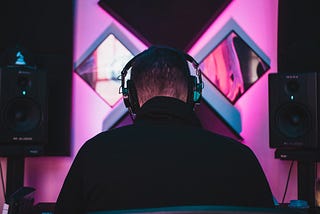 High contrast photo in blacks and neon pinks of the back of a person’s head and shoulders. They are wearing headphones and large audio equipment is visible in the background.