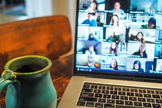 A green cup filled with coffee sits on a kitchen table next to a laptop running video conferencing software.