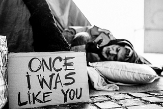 A homeless man sleeps on the street with a sign saying “Once I was like you.”