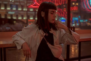 A woman is standing in front of neon lit stores on a rainy night.