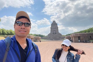 A smiling Asian man in a black sunglasses and beige cap, with black shirt and a blue sweater taking a wefie of himself with a smiling Asian lady in a white cap and aviator sunglasses, with a beige shirt and denim jacket. The lady has long hair and is picture grabbing a bunch of it with her left hand showing that she’s not ready for the shot. The background is the Monument to the Battle of the Nations in Leipzig, Germany — on a sunny day.