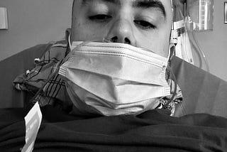 A black and white photo of a man in an ER room. He is in pain, hooked up to oxygen, wearing a surgical mask and is having a migraine attack. Credit: Joseph Coe
