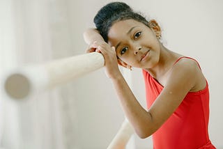 Girl in red leotard leaning on ballet barre, staring at camera