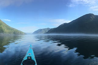 A panorama centred around the nose of a kayak, steep mountains on either side and an ocean channel with low fog in centre