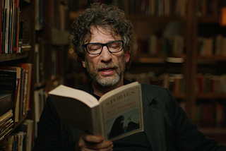 Neil Gaiman reading from one of his short stories to audience. He is wearing Black t-shirt with black suit.