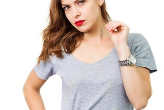 An attractive brunette in a light grey shirt stands with one hand on her hip while the other hand plays with the silver chain of her necklace as she stares at the camera.