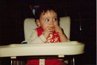 a baby sits in a retro-looking highchair with a party hat on and she’s biting into an orange segment with apparent trepidation