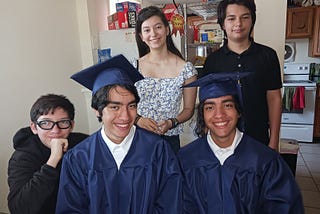 Five teenagers, one female, and four males. Two of the males are wearing navy blue caps and gowns in preparing for graduation.