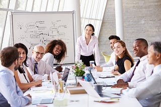 Asian Businesswoman Leading Meeting At Boardroom Table with other leaders, colleagues, and or peers of mixed races white, South-Asian, and Black all of different ages and work experience.