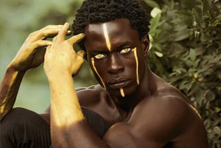 A man of African descent stares into the came lens wearing neon yellow contact lenses, four vertical yellow stripes of paint on his face, and his forearms are covered in yellow paint. He’s posing amidst bushes and greenery in the background.