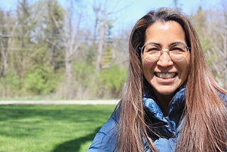 Photo of candidate Maria Peterson, running for IL House of Representatives, District 52. Maria is wearing a blue jacket, standing outside with green grass and trees in the background.