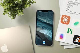 An iPhone 14 Pro on a table showing an empty home screen with a Safari icon. Next to it is a gold MacBook, a green plant and some papers. The top paper shows the text Apple Vision Pro, Apple Watch, iPhone, iPad , Mac. There are app five icons beneath the text.