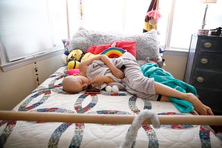 A young girl with cancer, lays on her bed looking a little nervous, surrounded by pillows and stuffed animals.