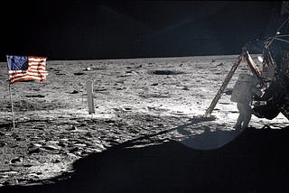 An astronaut stands by the lunar module to the right. An American flag appears on the left on the surface of the moon.