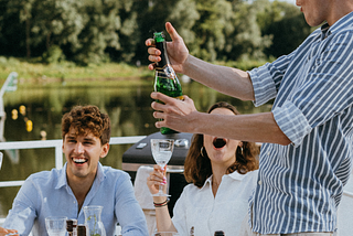 A man in a striped blue and white shirt pops open a bottle of champagne. Behind him a woman in a white shirt is holding a glass and looking up at him. Next to her is a man wearing a blue shirt and laughing. They are sat outside by a lake.