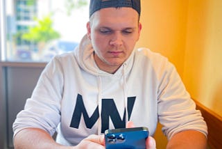 Isaac Mashman wearing a backward hat and white Mashman Ventures hoodie, sitting in front of an orange wall, looking at his phone.